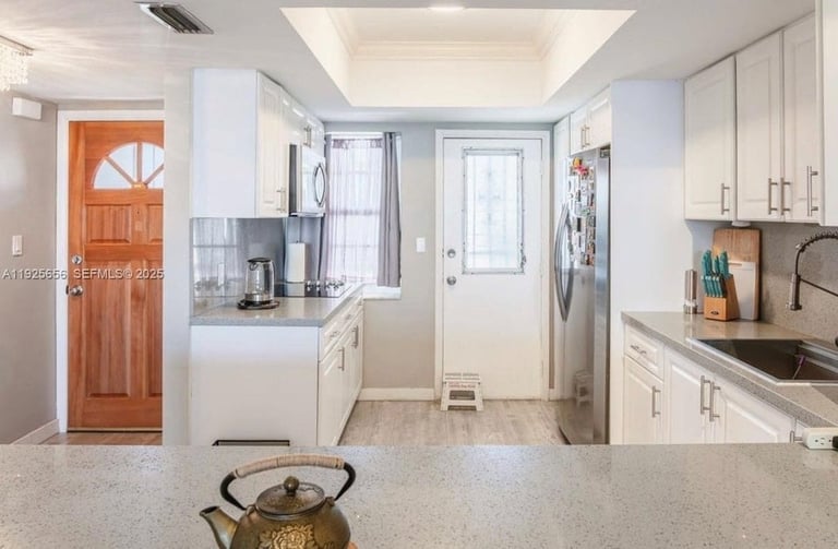 Bright modern kitchen with white cabinetry, stainless steel appliances, and a pass-through to living area with wooden entry door